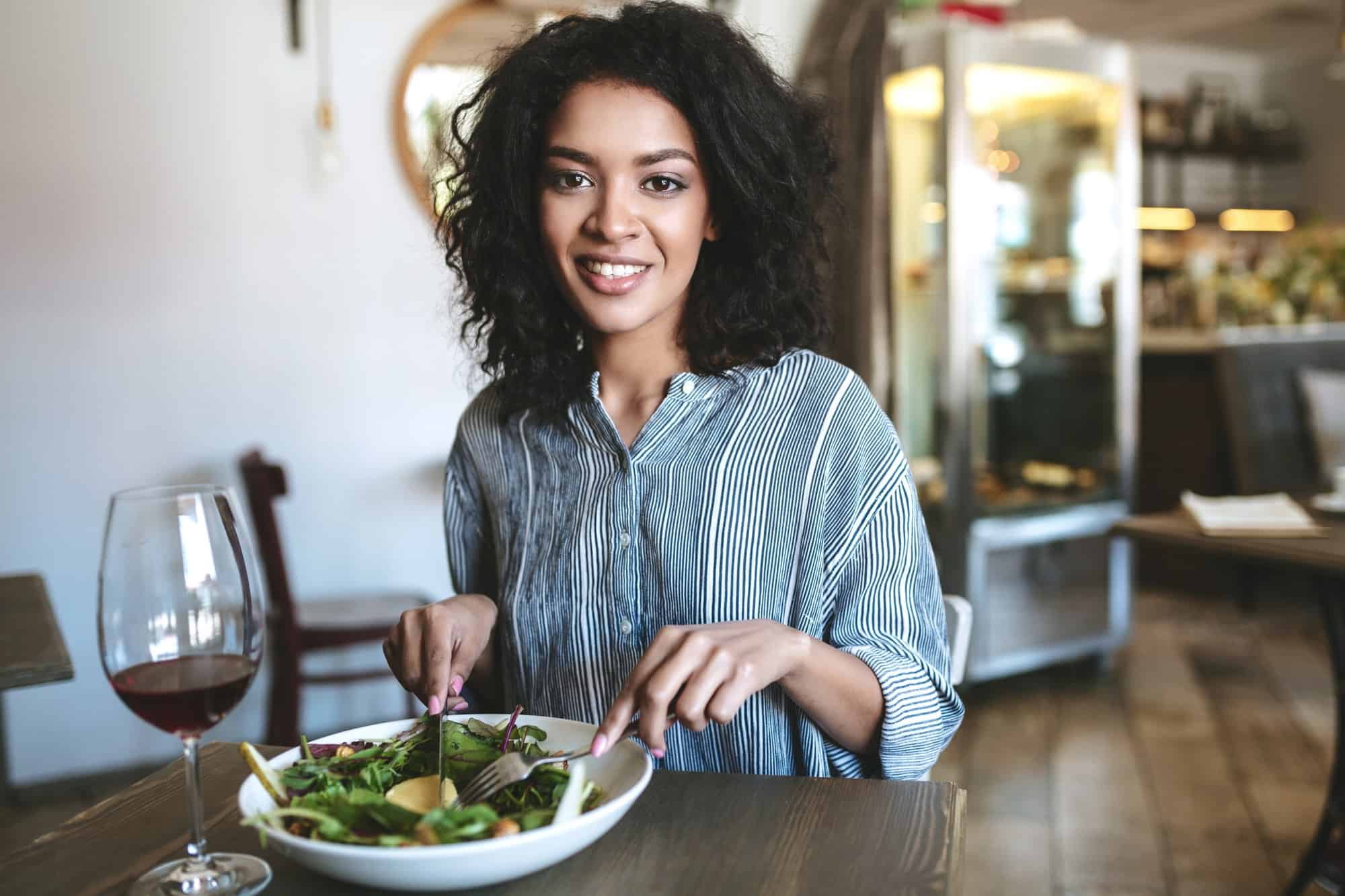 Smiling girl looking in camera with glass of red wine and salad on table at cafe