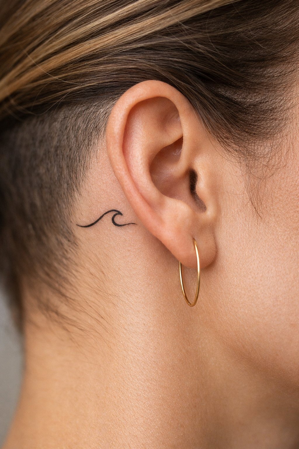 Behind-the-ear macro: a minimalist wave line, small and sleek. The hair is shaved slightly to showcase it, and a delicate gold earring is in the frame.