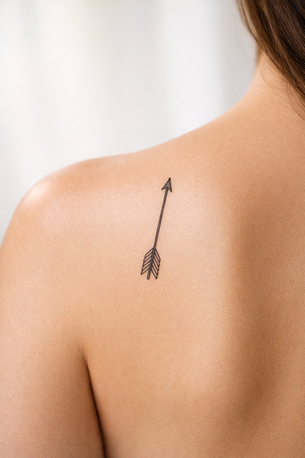Diagonal shot on the shoulder blade: a single, thin arrow tattoo, pointing upward. Clean lines, with the fletching (feathers) delicately detailed.