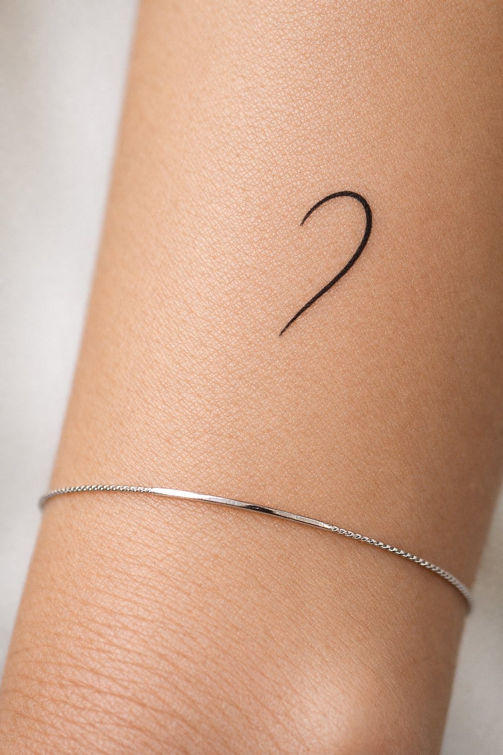 Macro photograph of an'open' heart tattoo on the inner forearm—only the curved right side is drawn. A slender, simple bracelet sits an inch below it on the wrist. The composition emphasizes the negative space.