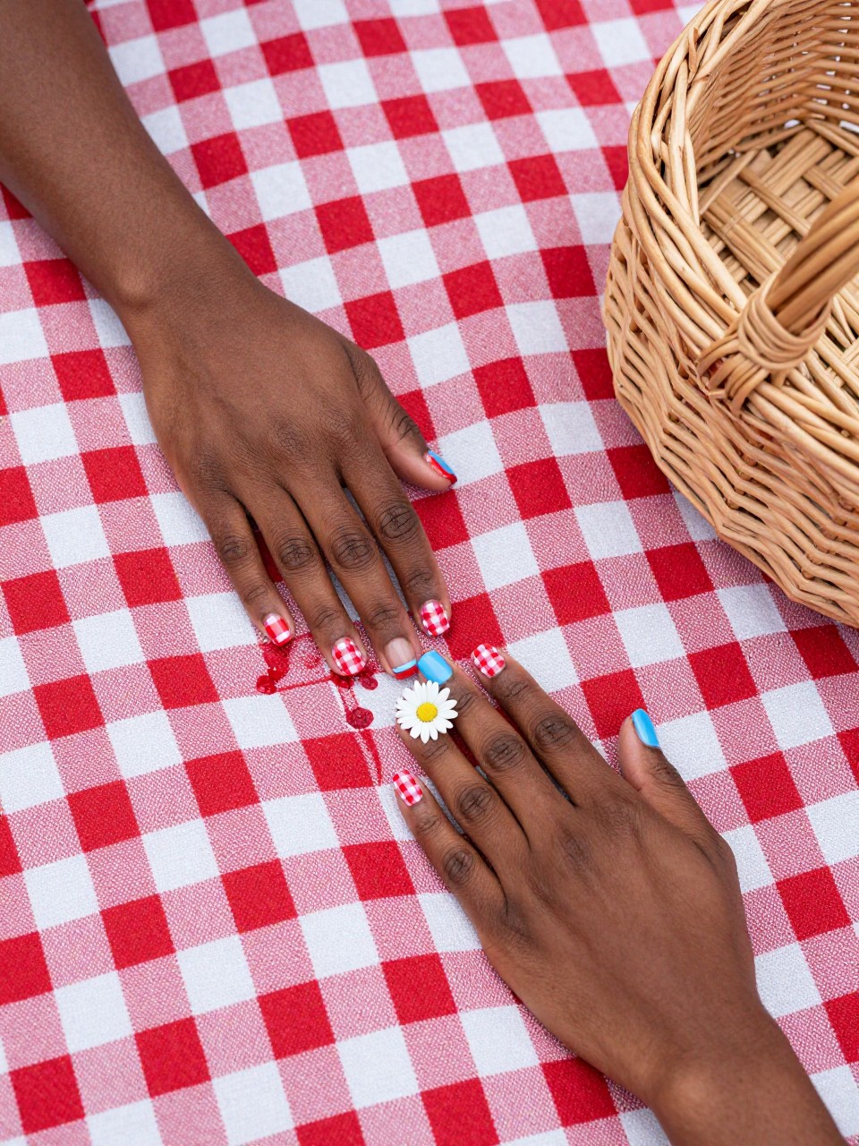 Top-down view of black female hands with short nails on a classic red gingham picnic blanket. Some nails are red and white gingham. The accent nails are baby blue with one bold white daisy. A wicker basket sits nearby in the grass.