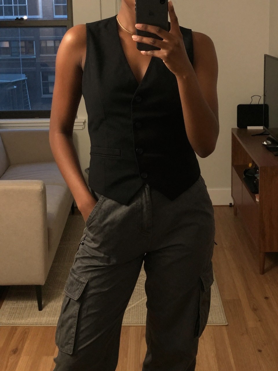 iPhone mirror selfie of a black woman wearing a black vest with cargo pants, posed with one hand in her pocket and phone in the other, cropped at NECK so face doesn't show, in a city apartment, warm moody lighting, iPhone photo quality.