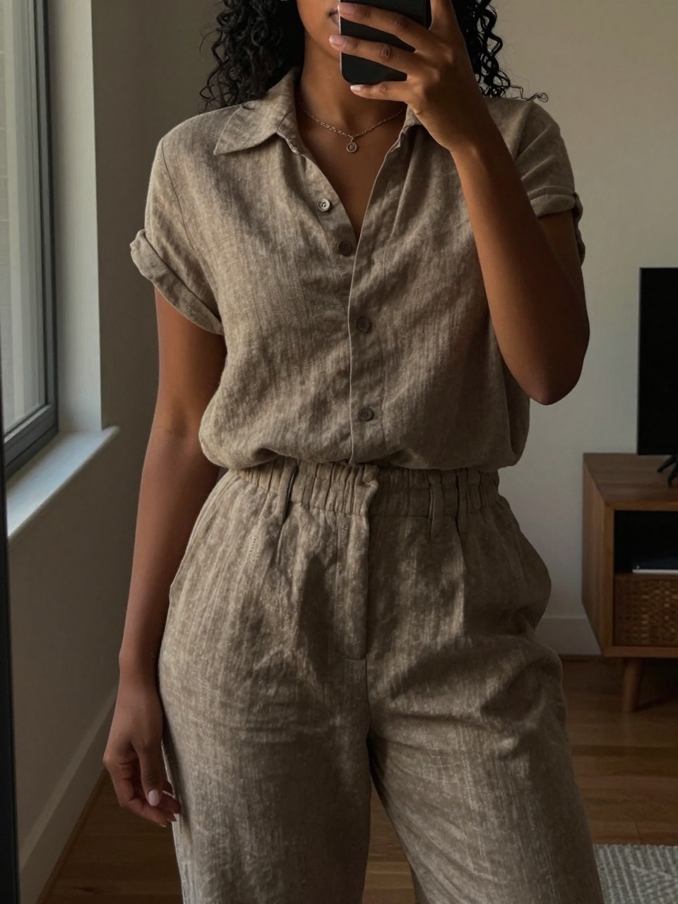 iPhone mirror selfie of a black woman wearing a taupe linen matching set, posed with her arms slightly relaxed, cropped at NECK so face doesn't show, in a city apartment, warm moody lighting, iPhone photo quality.