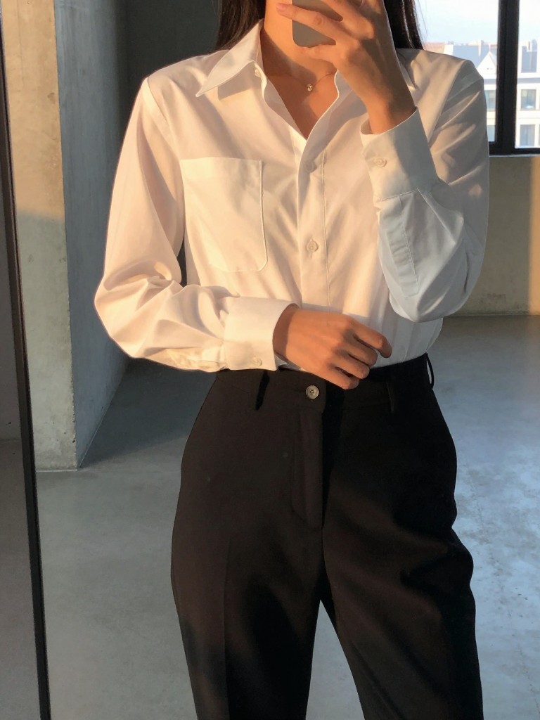 mirror selfie of a woman in a white blouse and black pants, adjusting her sleeve, cropped at neck so face doesn't show, in a modern loft with concrete walls, evening golden hour, dslr photo quality.
