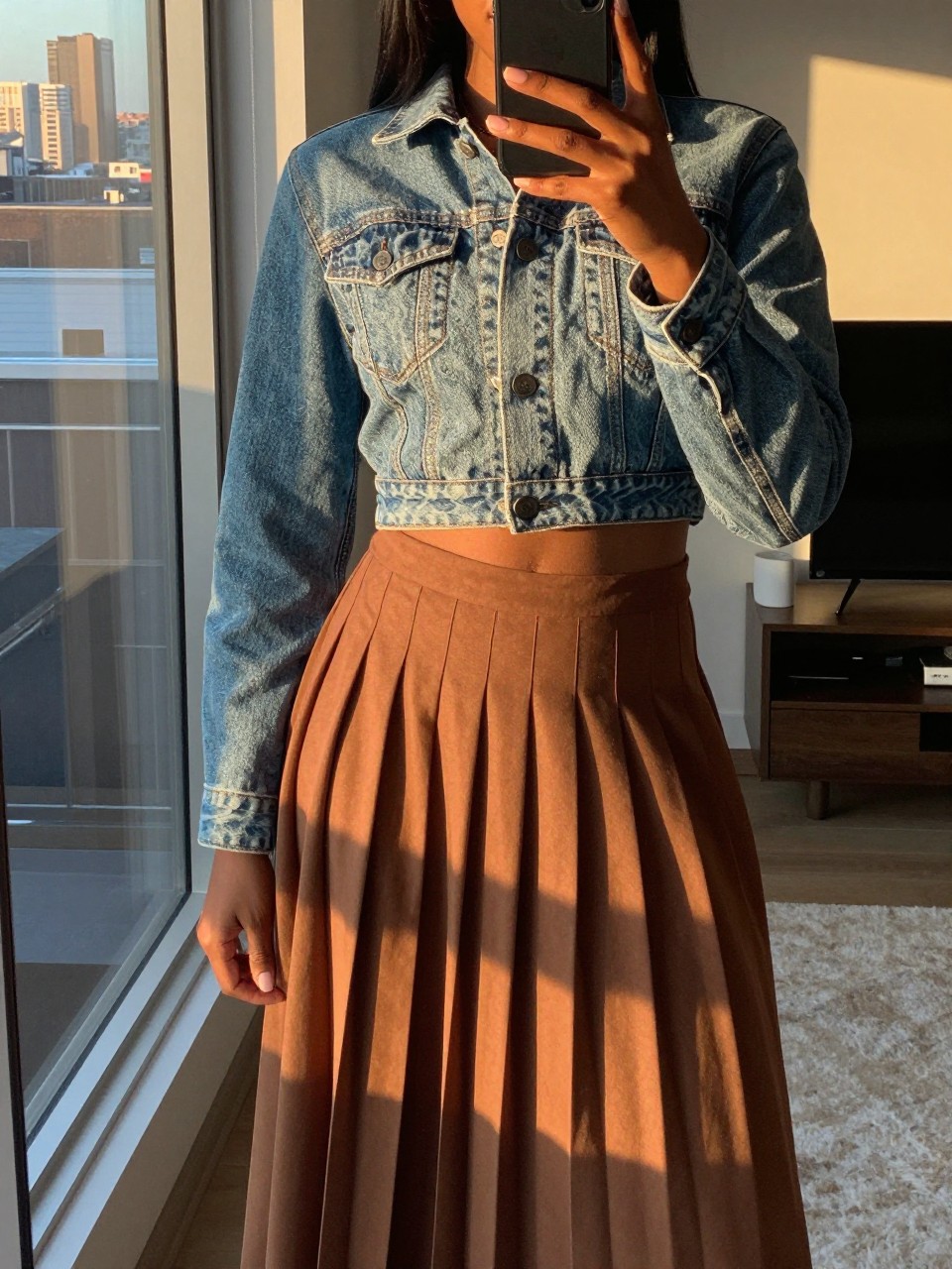 mirror selfie of a black woman in a cropped jean jacket and pleated midi skirt, phone held low to show skirt, cropped at NECK so face doesn't show, in a city apartment during evening golden hour, iPhone photo quality.