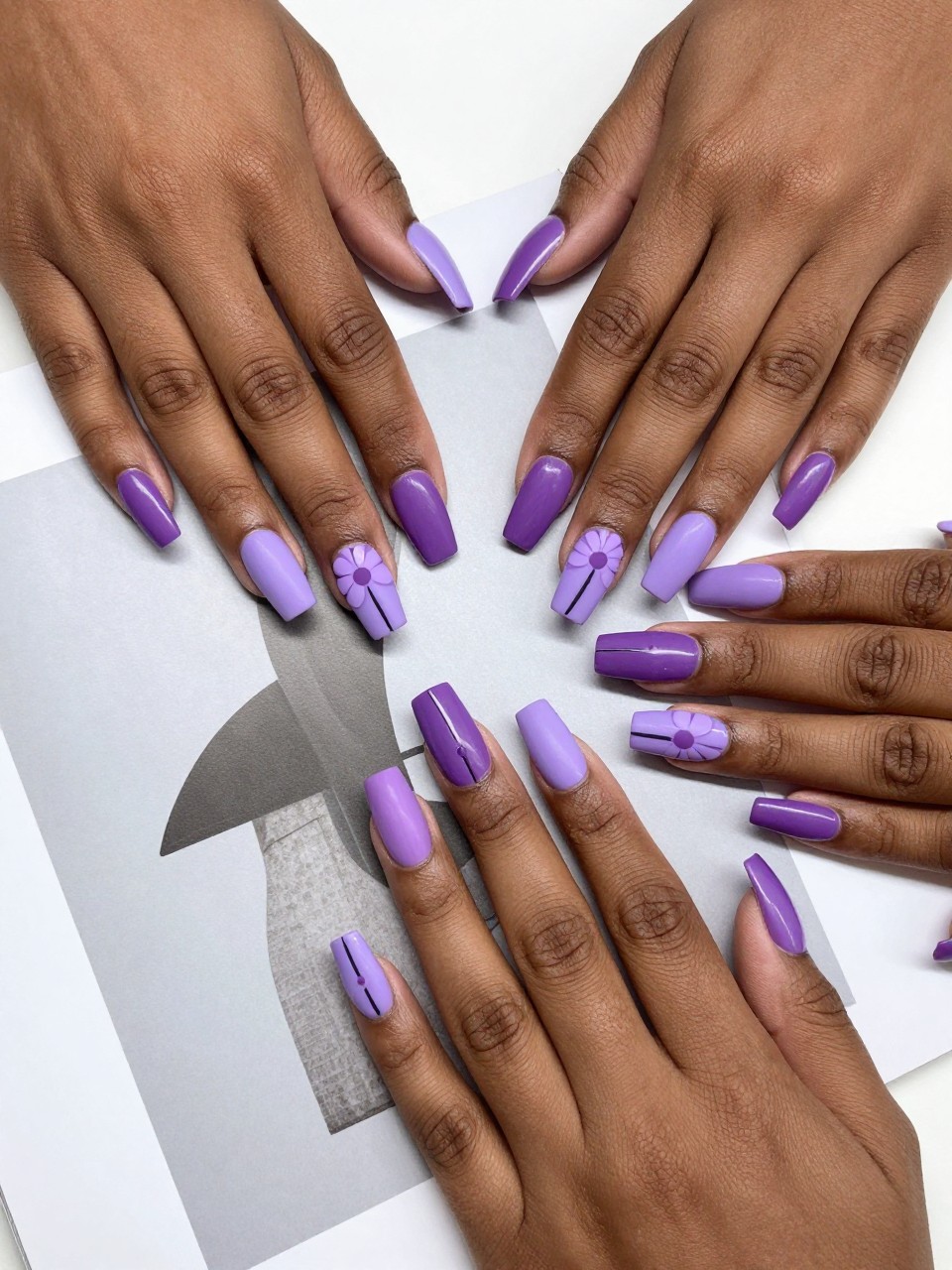 Overhead shot of black feminine hands with coffin designed nails laid on a modern art magazine. Each nail design is split diagonally: one half lilac, the other half plum. A simple, graphic flower in a dusty purple is painted at the seam. The look is crisp and tonal.