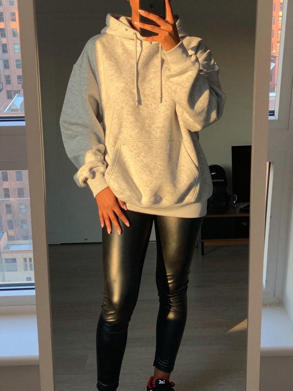 iPhone mirror selfie of a black woman in an oversized hoodie and leather leggings with Timbs, relaxed pose, cropped at NECK so face doesn't show, in a city apartment during evening golden hour, iPhone photo quality.
