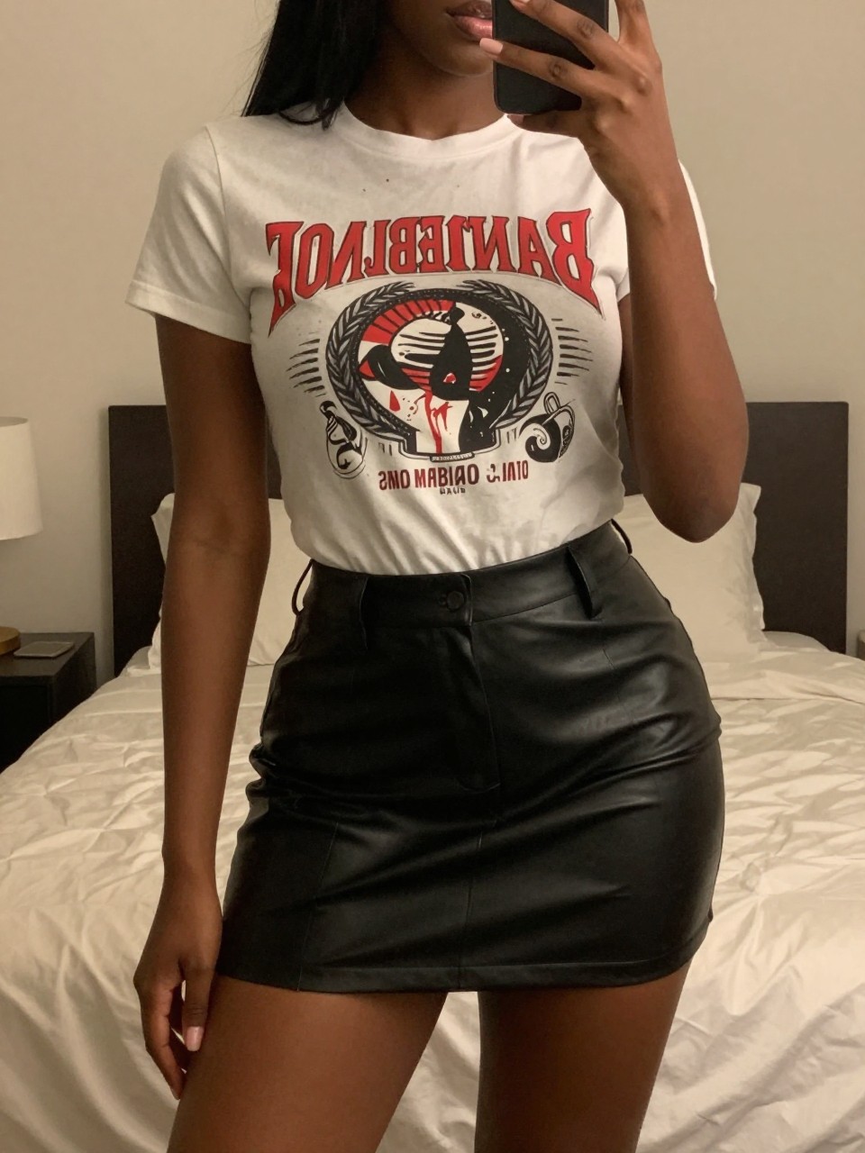 mirror selfie of a black woman in a graphic tee and leather skirt, relaxed pose with slight turn, cropped at NECK so face doesn't show, in a feminine-themed bedroom with warm moody lighting, iPhone photo quality.