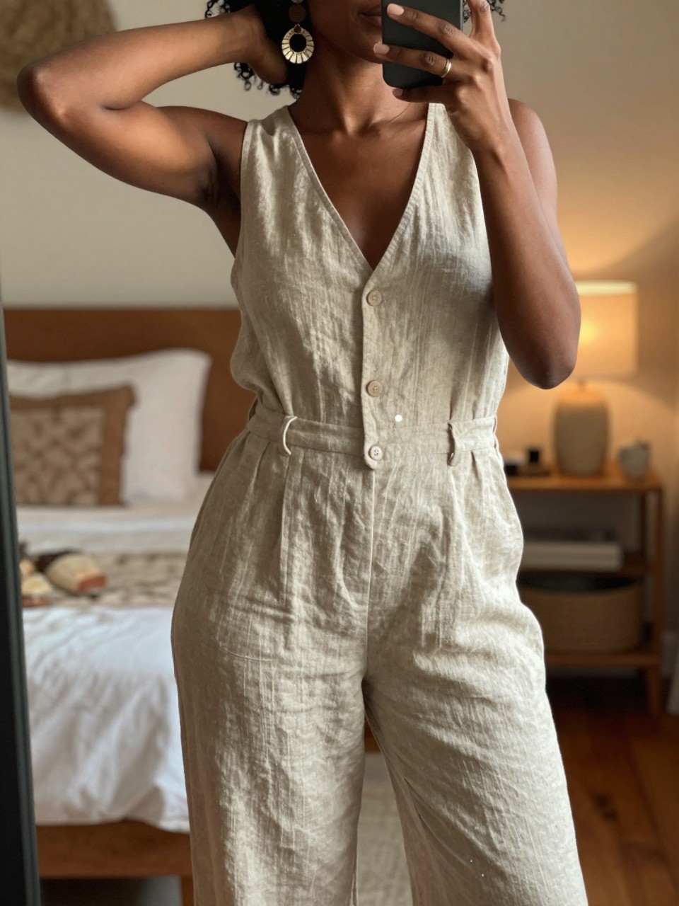 iPhone mirror selfie of a black woman in a natural linen jumpsuit and statement earrings, one hand behind head, cropped at NECK so face doesn't show, in a bohemian-themed bedroom with warm moody lighting, iPhone photo quality.