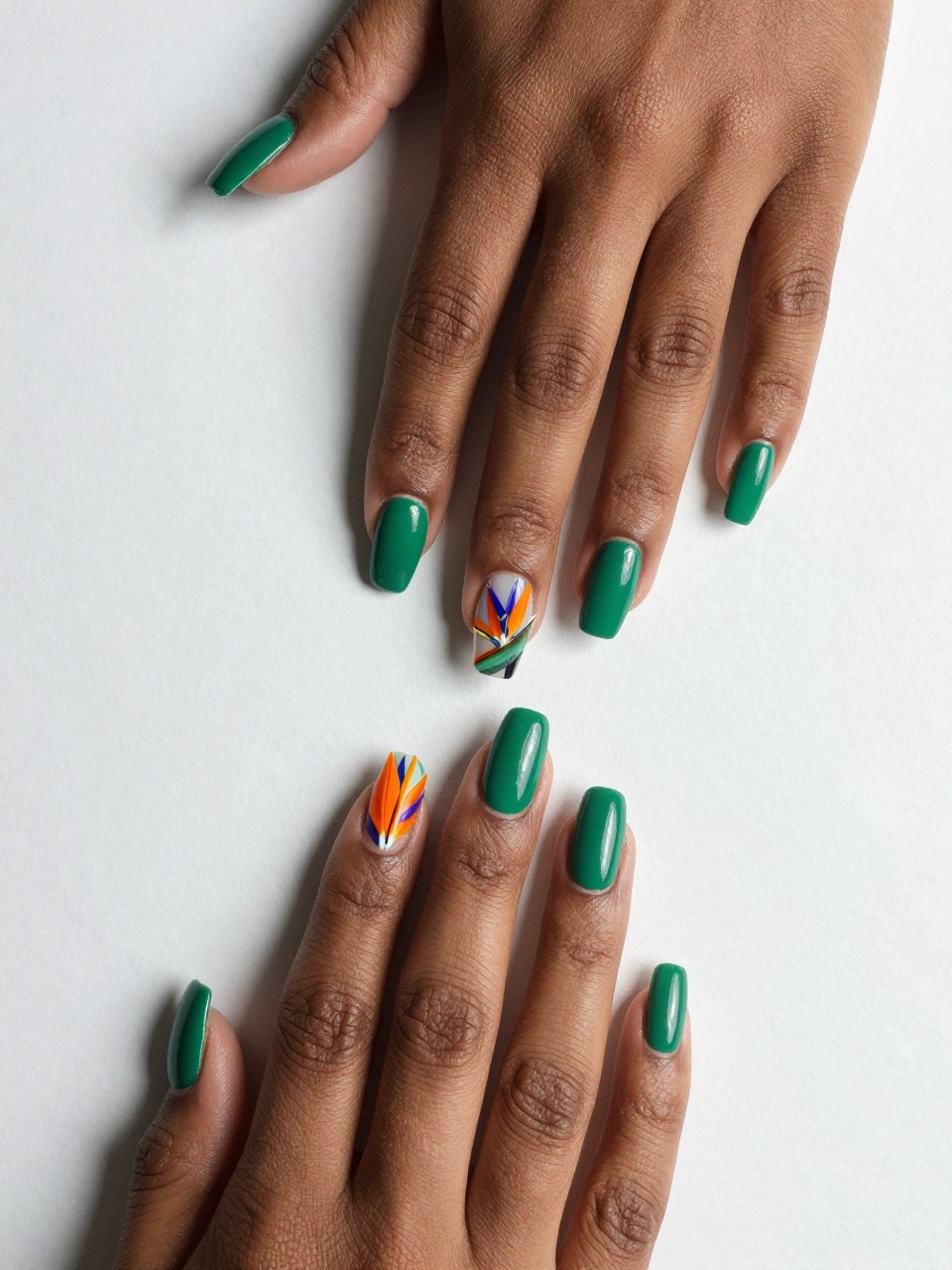 Overhead shot of black female hands with coffin nails, one hand resting on a table. The nails are glossy emerald green. The accent nails feature a large, detailed Bird of Paradise bloom in bright orange and purple-blue.