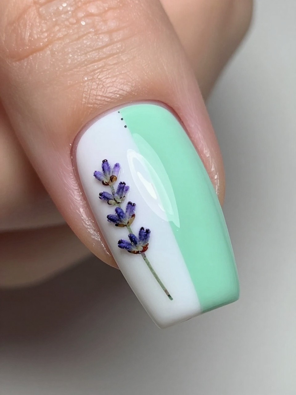 Angled overhead view of a nail. The French tip is split vertically: half white, half mint green. From the seam in the middle, a small painted sprig of lavender extends down the side of the nude nail bed.