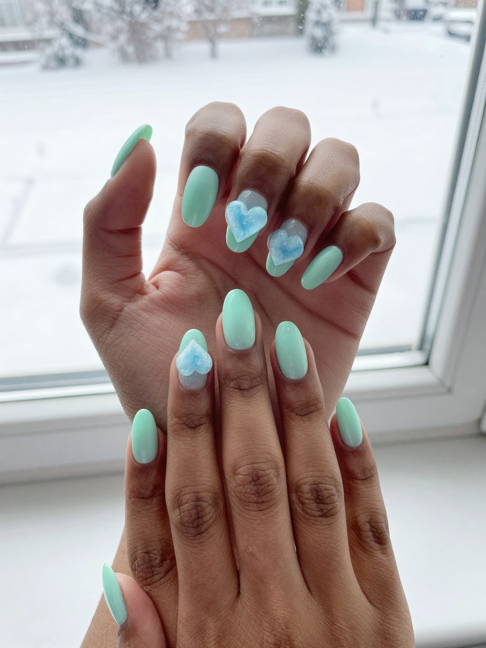 Bird's-eye view of black feminine hands against a snowy windowsill backdrop. The nails are a frosted mint green. A frosted ice-blue heart is centered on each nail. The overall feel is chilly, sweet, and cohesive.