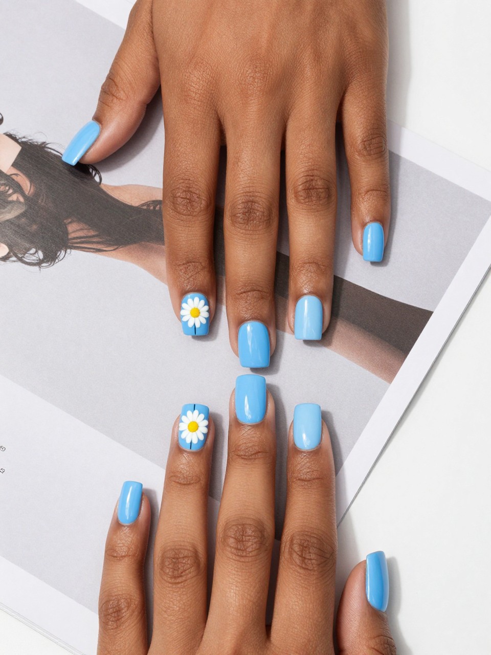 Top-down view of black feminine hands with square nails on a modern design magazine. Each nail is split diagonally: one half powder blue, the other half sky blue. A simple, geometric white daisy is painted at the seam. The look is crisp and bright.