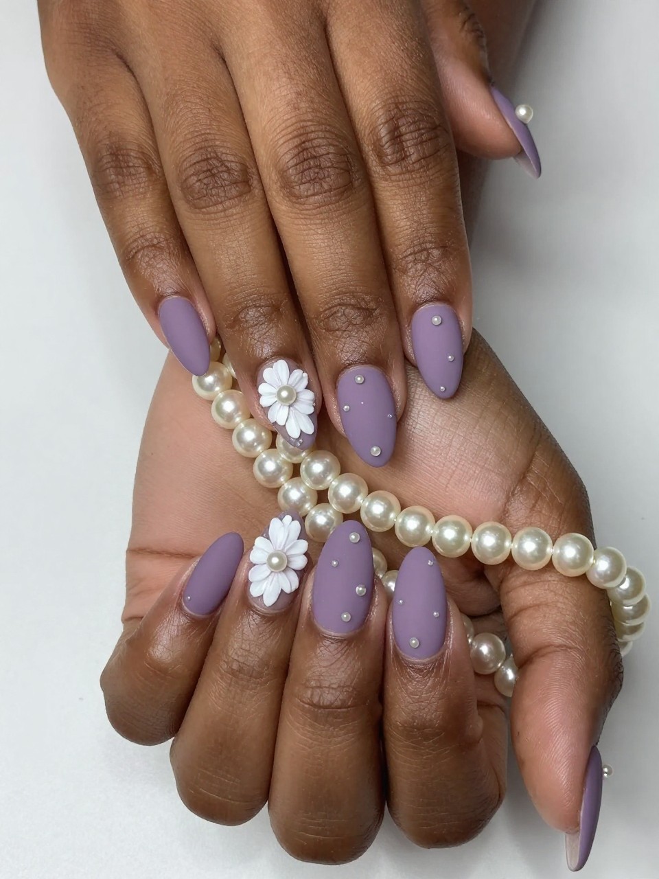 Overhead view of black feminine hands with almond nails draped over a strand of pearls. The nails have a matte dusty purple base. Small white flowers dot each nail, each with a single micro pearl in the center. Soft lighting highlights the tiny raised textures.