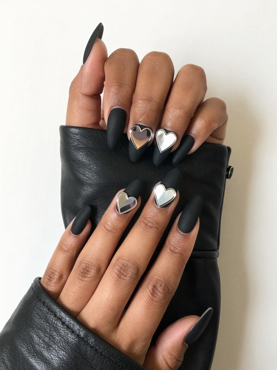 Overhead, top-down view of black feminine hands with long, coffin-shaped nails resting on a black leather jacket sleeve. The nails are a flat matte black. On each nail, a single, perfect mirror-chrome heart is placed off-center. Sharp, studio lighting reflects brilliantly in the chrome. The vibe is rock-and-roll chic.