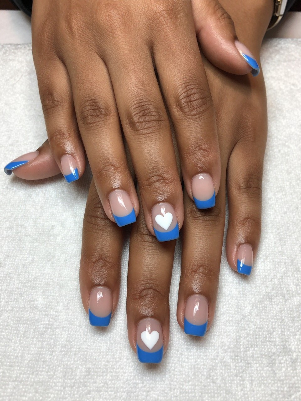 Close-up of black feminine hands with a baby blue French manicure. A single micro white heart is painted right on the line of each blue tip. The hands rest on a clean white towel, looking pristine and subtly detailed.