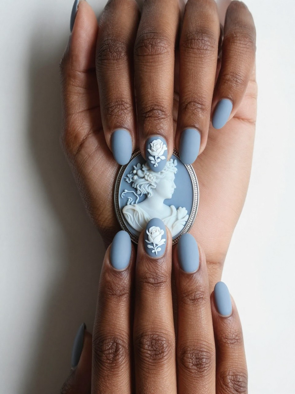 Overhead view of black feminine hands with oval nails resting on a blue and white cameo brooch. The nails are a matte Wedgwood blue. A raised, white floral silhouette (like a rose profile) is painted in the center of each nail. The look is heirloom-elegant.