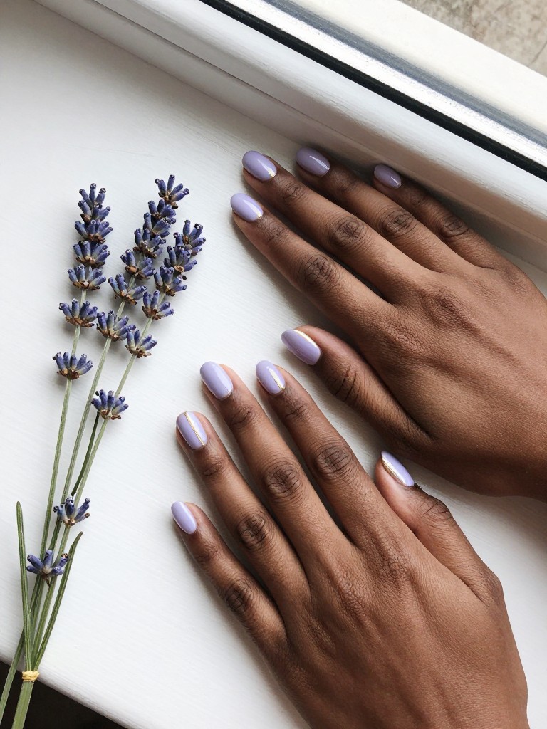 Overhead view of black hands resting beside a sprig of lavender. The nails are short and oval, painted a milky pastel lavender. A delicate, vertical line of metallic gold runs along the outer side edge of each nail. The setting is a bright, white windowsill with clean light.
