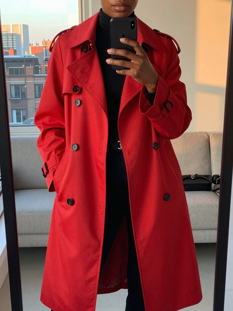 iPhone mirror selfie of a black woman in a red trench coat over all-black, hand in coat pocket, cropped at NECK so face doesn't show, in a city apartment during evening golden hour, iPhone photo quality.