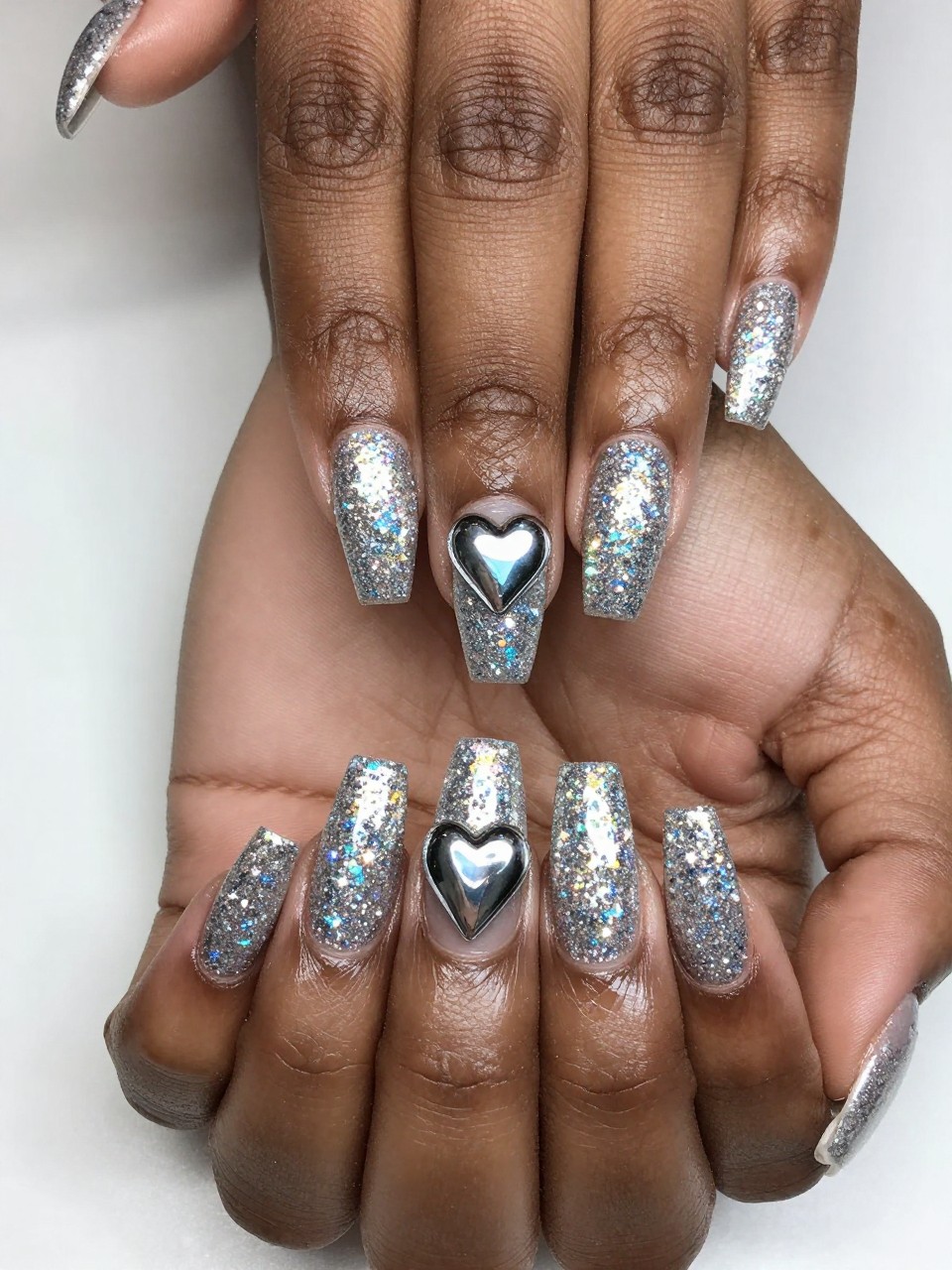 Overhead shot of black feminine hands under bright light, showcasing extreme sparkle. The nails are densely covered in holographic silver glitter. A single, perfect chrome heart sits in the center of each nail, reflecting the light differently than the glitter around it.