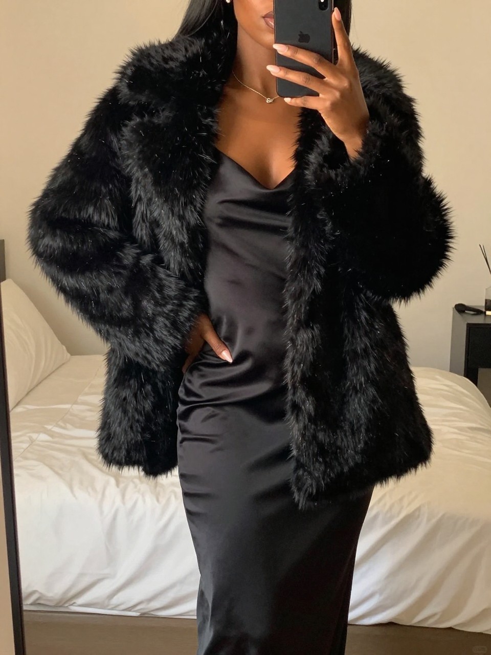 iPhone mirror selfie of a Black woman wearing a black faux fur coat over a satin slip dress, posed with one hand on her hip and phone in the other, cropped at NECK so face doesn't show, in a feminine-themed bedroom, evening golden hour light, iPhone photo quality.