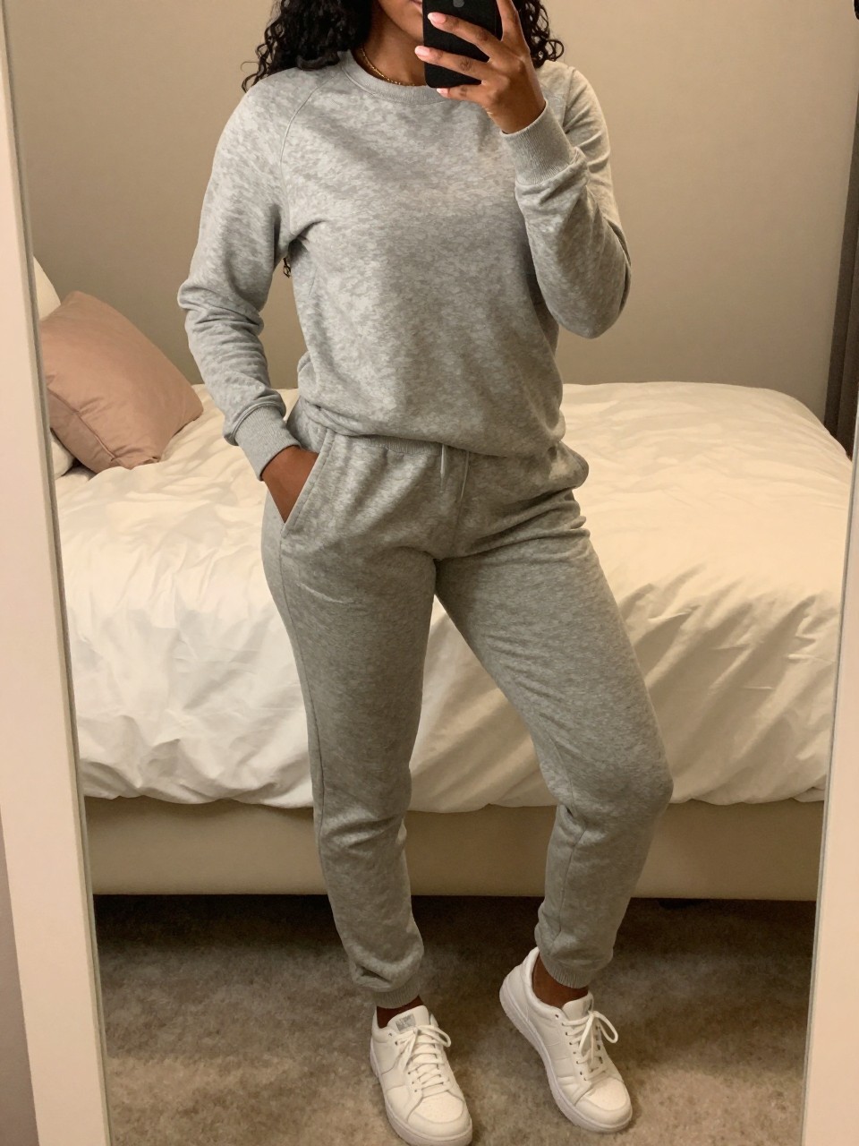 iPhone mirror selfie of a black woman in a grey matching sweatsuit and white sneakers, relaxed pose with one hand in pocket, cropped at NECK so face doesn't show, in a feminine-themed bedroom with warm moody lighting, iPhone photo quality.