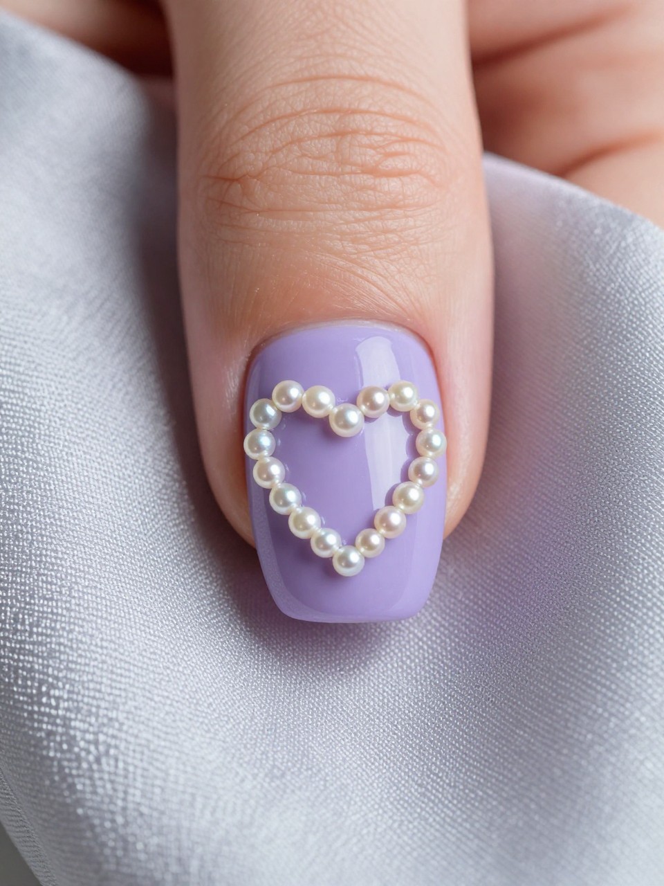 Macro, top-down view of a nail. The base is a soft periwinkle. A heart shape is traced with tiny, iridescent pearls. The hand rests on a silk scarf, with the pearls catching the light delicately.