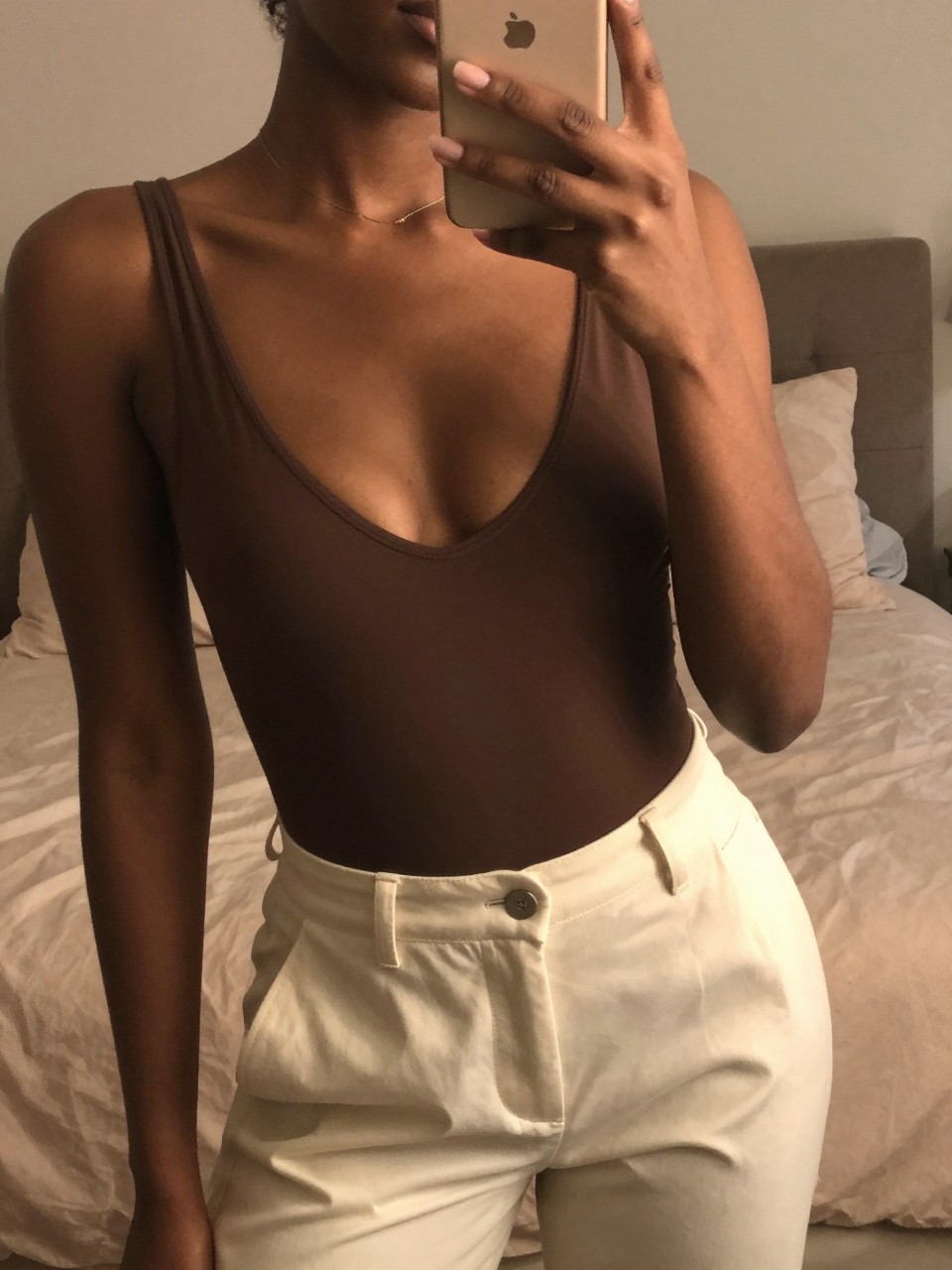 iPhone mirror selfie of a black woman in a plunge bodysuit and cream trousers, angled slightly, cropped at NECK so face doesn't show, in a feminine-themed bedroom with warm moody lighting, iPhone photo quality.