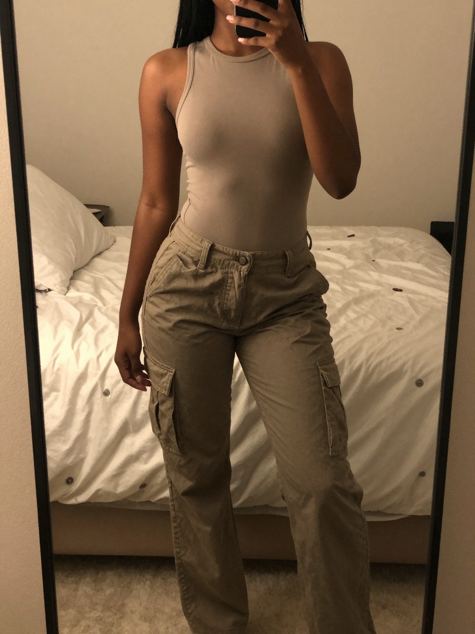 iPhone mirror selfie of a black woman in a mock neck bodysuit and cargo pants, one foot slightly forward, cropped at NECK so face doesn't show, in a feminine-themed bedroom with warm moody lighting, iPhone photo quality.