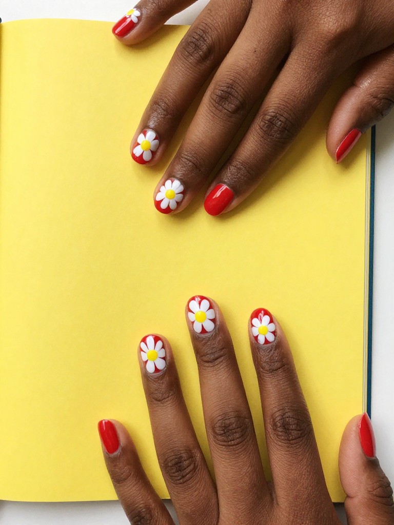 Overhead view of black hands with short nails on a bright yellow notebook. The nails are glossy fire-engine red. Simple, cartoonish white daisies with big yellow centers are painted on each nail. The vibe is energetic and fun.