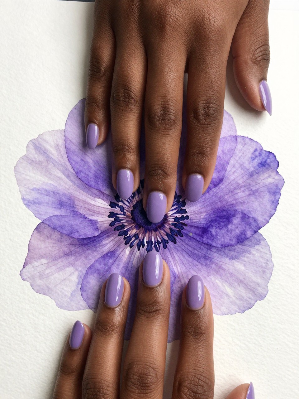 Close-up, top-down view of black feminine hands with oval nails on watercolor paper. The nails have a soft, blurred wash of lilac. A single, more detailed purple anemone with a dark center is painted clearly on top of the wash. The style is artistic and layered.