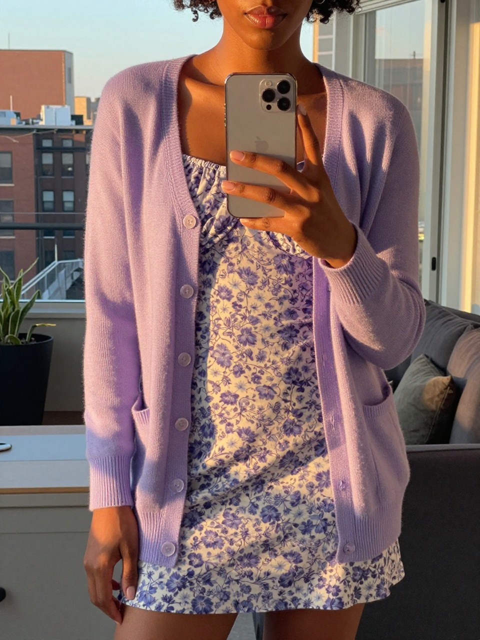 iPhone mirror selfie of a Black woman wearing a lavender cardigan over a floral dress, standing casually with her phone low, cropped at NECK so face doesn't show, in a city apartment, evening golden hour light, iPhone photo quality.