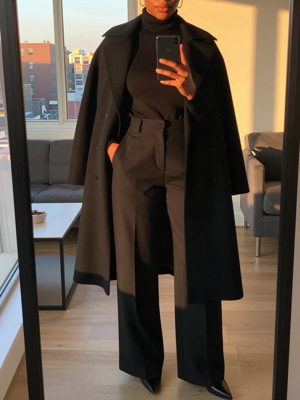 iPhone mirror selfie of a Black woman wearing a black cape coat over a turtleneck and wide-leg pants, posed with one hand in her pocket and phone in the other, cropped at NECK so face doesn't show, in a city apartment, evening golden hour light, iPhone photo quality.
