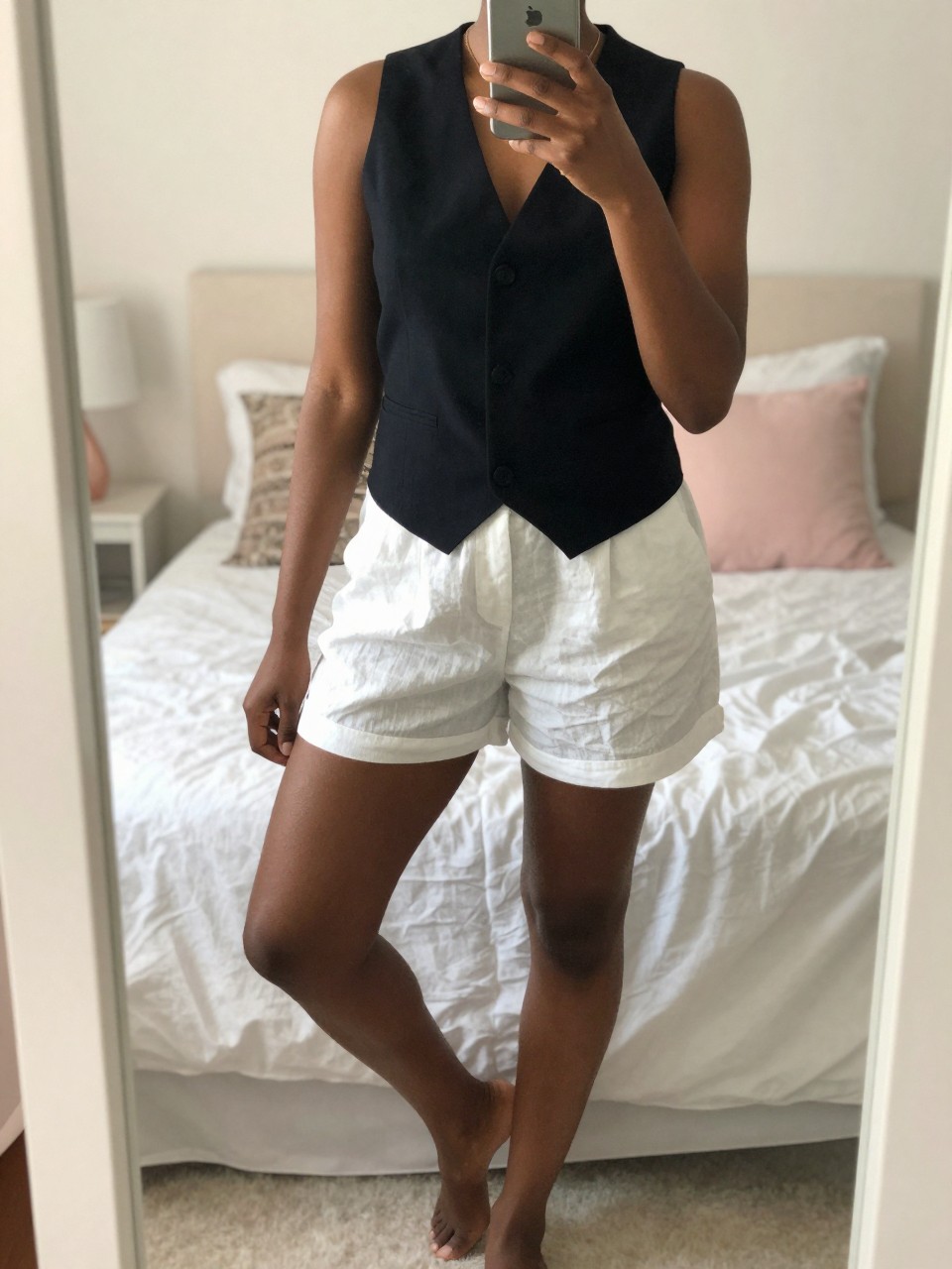 iPhone mirror selfie of a black woman wearing a black vest with white linen shorts, standing casually with one foot forward, cropped at NECK so face doesn't show, in a bright feminine-themed bedroom, warm moody lighting, iPhone photo quality.