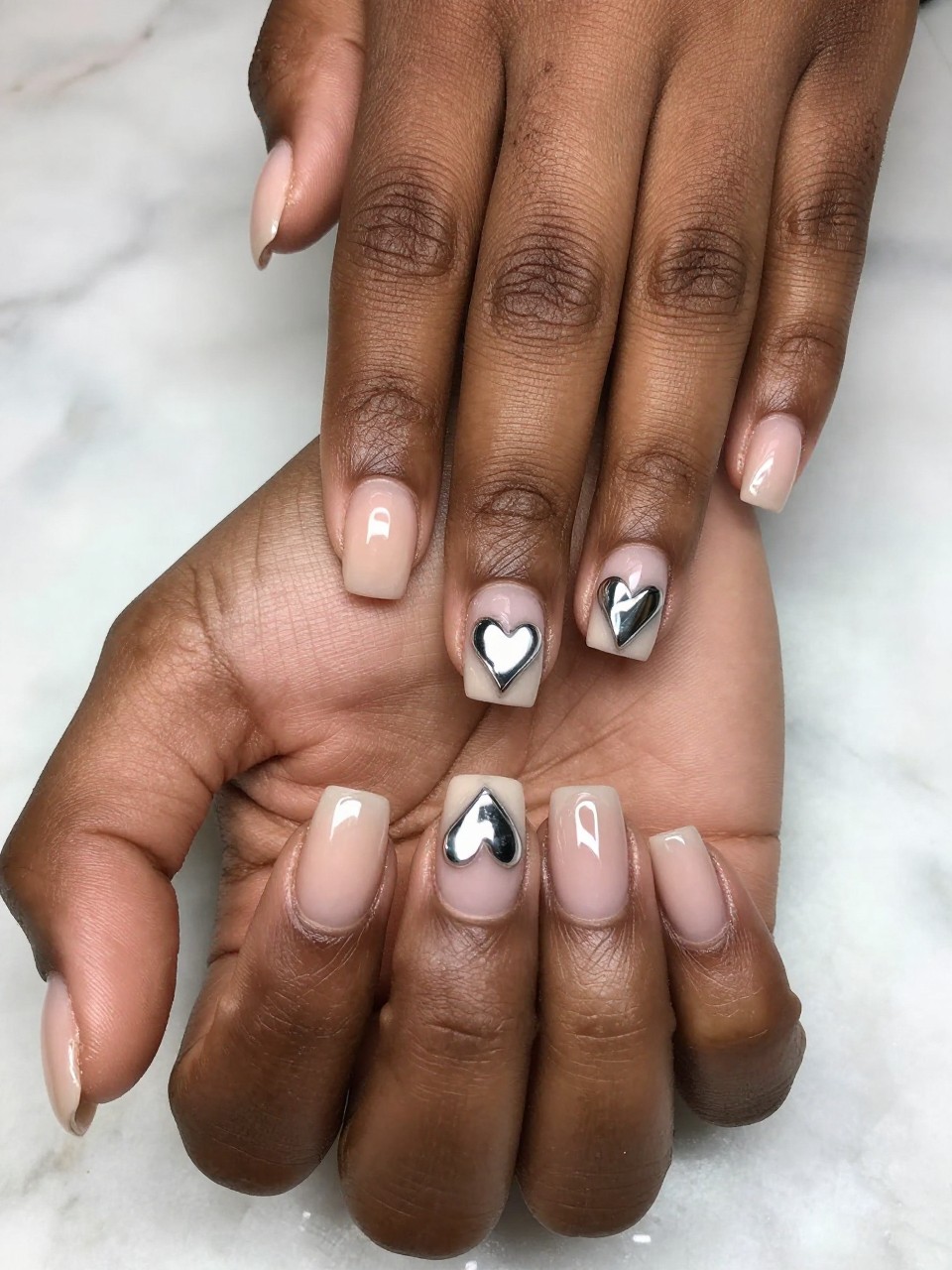 Close-up, top-down view of black feminine hands with short, squared nails on a marble surface. The nails have a sheer nude base. A perfect heart shape in the center of each nail reveals a layer of bright mirror chrome beneath. The look is clean, precise, and futuristic.