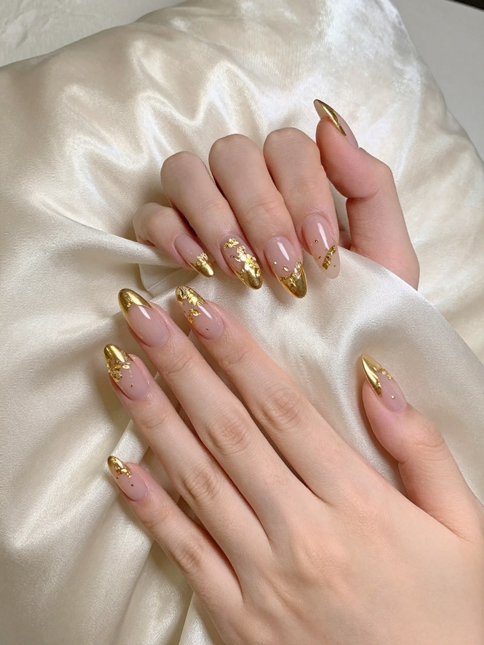Bird's-eye view of hands with almond nails draped over a silk pillow. The nails have a shiny gold French tip. On the sheer pink base, delicate flakes of gold leaf are placed in a loose floral pattern. The lighting is warm and luxurious.