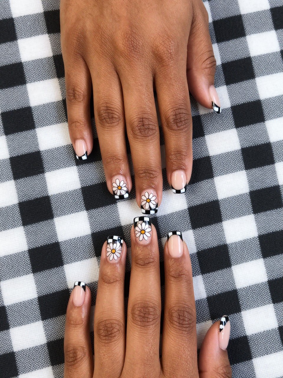 Top-down view of black feminine hands with short, square nails on a gingham blanket. The French tips are a neat black and white check. On the nude base of each nail, a simple daisy outline in black is painted. The look is trendy and cute.