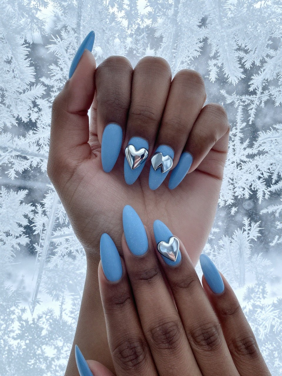 Overhead view of black feminine hands with long, almond nails against a backdrop of icy window frost. The nails are a matte icy blue. Frosted, silvery chrome hearts are placed on each nail. The lighting is cool and bright, creating a wintery vibe.