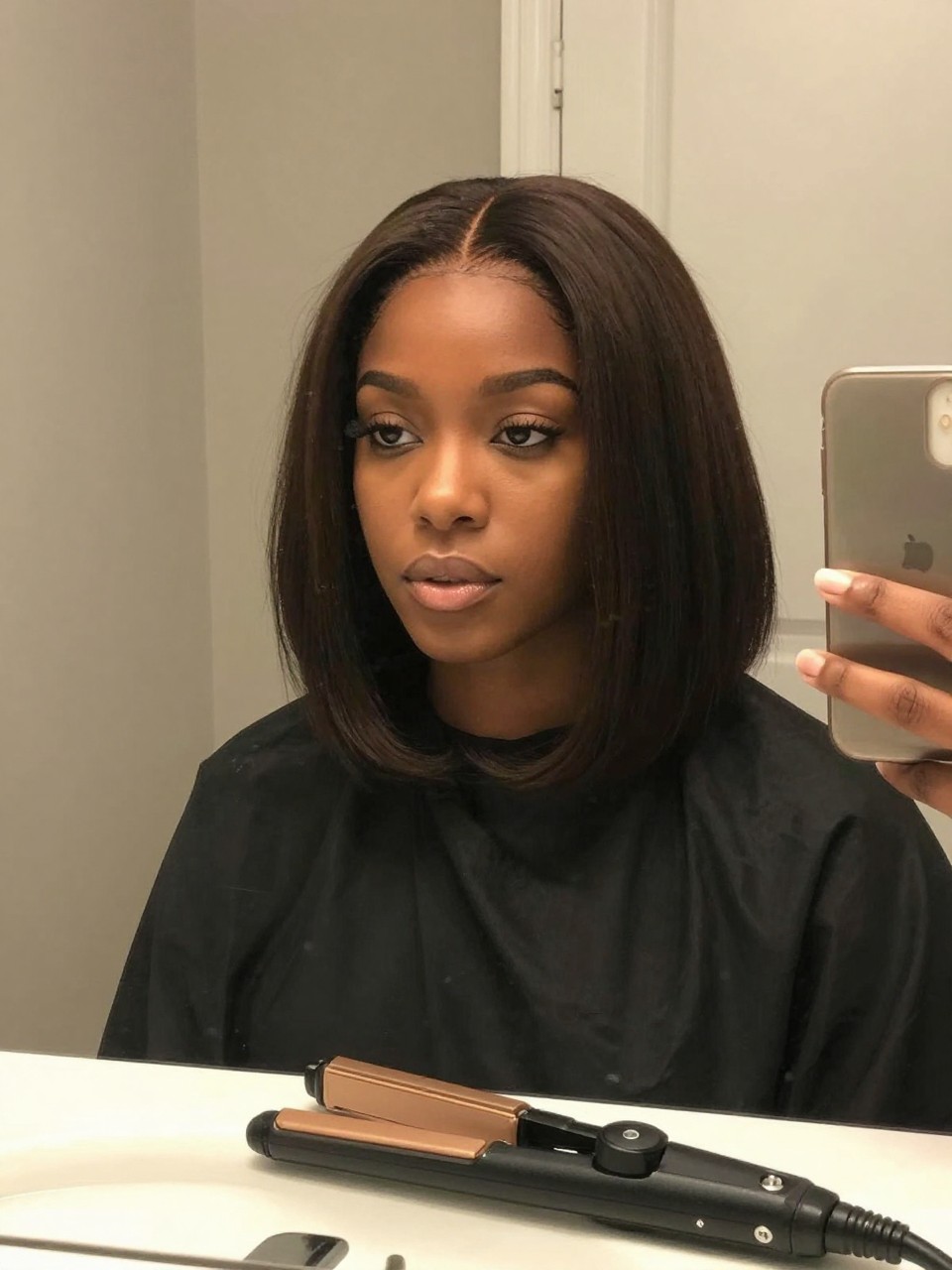 Photo of a black woman wearing a classic chin-length layered bob, front-facing view showing the layers framing her face, setting is a bathroom vanity mirror with soft indoor lighting, containing a flat iron resting on the counter, iPhone photo quality.