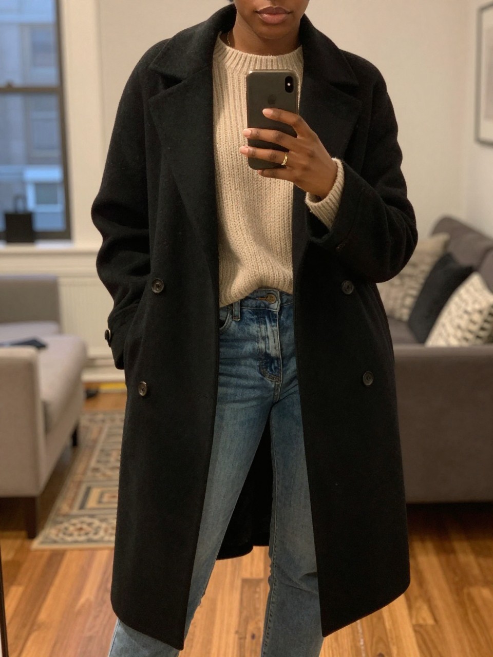 iPhone mirror selfie of a Black woman wearing a black cocoon coat over a chunky oatmeal sweater and jeans, posed casually with her phone low, cropped at NECK so face doesn't show, in a city apartment, warm moody lighting, iPhone photo quality.