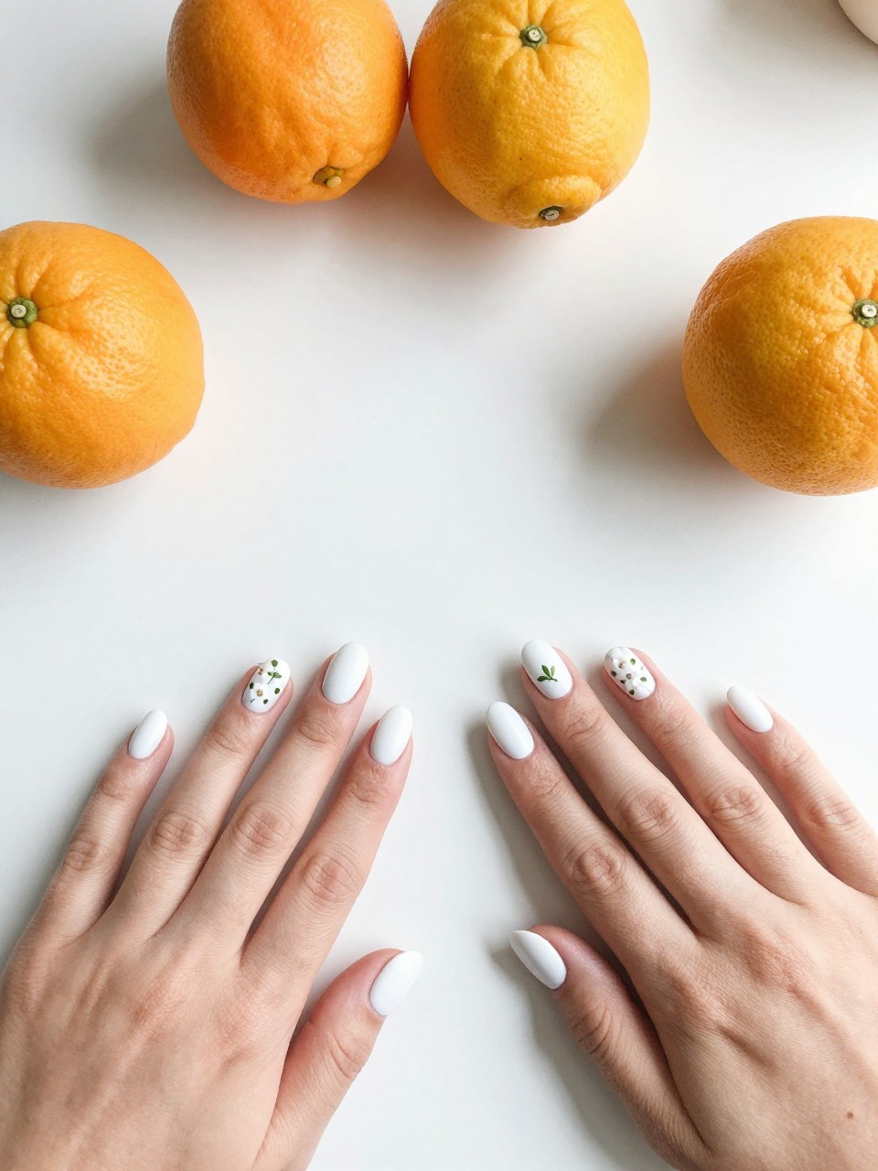 Top-down view of hands with oval nails on a white kitchen counter with citrus fruits. The nails are matte white. Delicate citrus blossoms and tiny green leaves are painted in a sparse, artistic arrangement on each nail. The light is bright and clean.