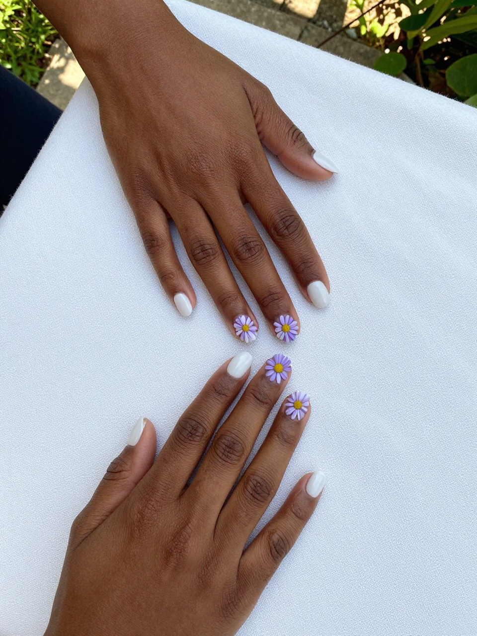 Overhead, top-down view of black feminine hands with short, rounded nails resting on a white linen tablecloth. The nails have a matte white base. Two or three lavender daisies with sunny yellow centers are painted on each nail. The setting is a bright, sunlit garden patio.