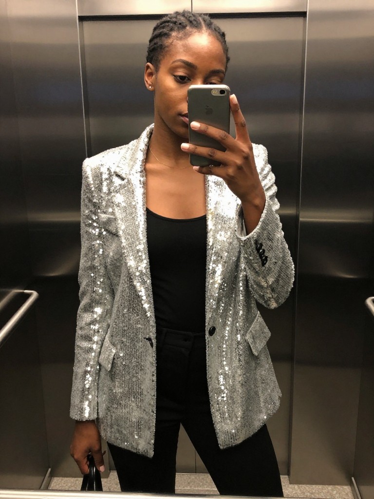 mirror selfie of a black woman wearing a silver sequin blazer over a black tank and trousers, blazer catching the light, cropped at torso, in a dark elevator with reflective walls, iPhone photo quality.