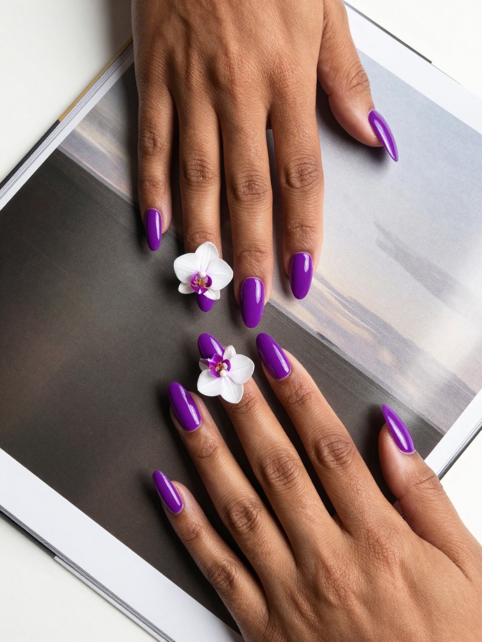 Overhead shot of black feminine hands with coffin nails resting on a glossy art book. The nails are a bright glossy grape purple. Each ring finger features a single, meticulously painted white orchid with graceful, clean lines. The lighting is bright and modern.