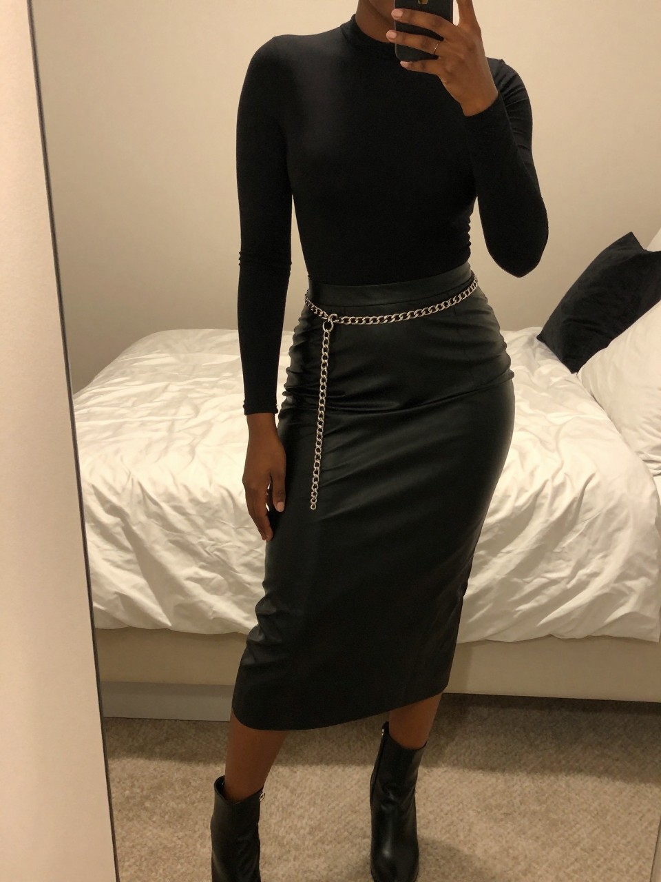 iPhone mirror selfie of a black woman wearing a black bodysuit tucked into a black leather midi skirt with a silver chain belt and black ankle boots, one hand resting on the belt, cropped at NECK so face doesn't show, in a feminine-themed bedroom, warm moody lighting, iPhone photo quality.