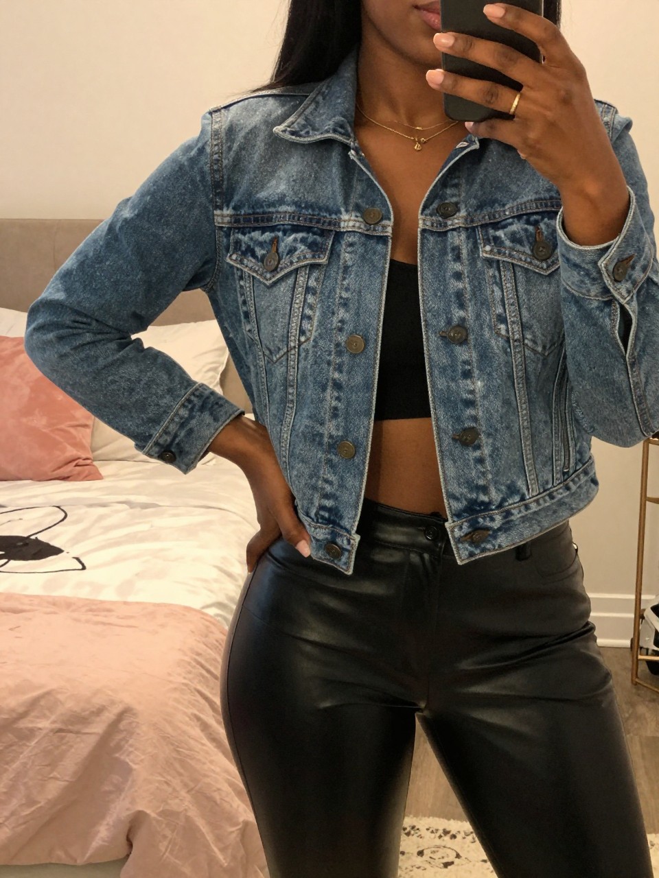 mirror selfie of a black woman in a jean jacket and leather pants, one hand on hip, cropped at NECK so face doesn't show, in a feminine-themed bedroom with warm moody lighting, iPhone photo quality.
