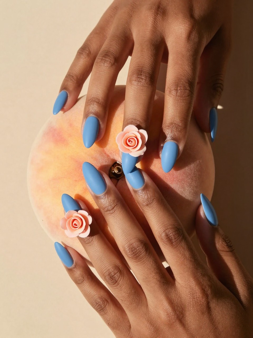 Overhead view of black feminine hands with almond nails lightly touching a peach. The nails are a matte powder blue. Each ring finger features a single, meticulously painted peach rose with subtle depth. The setting is warm and sunlit.