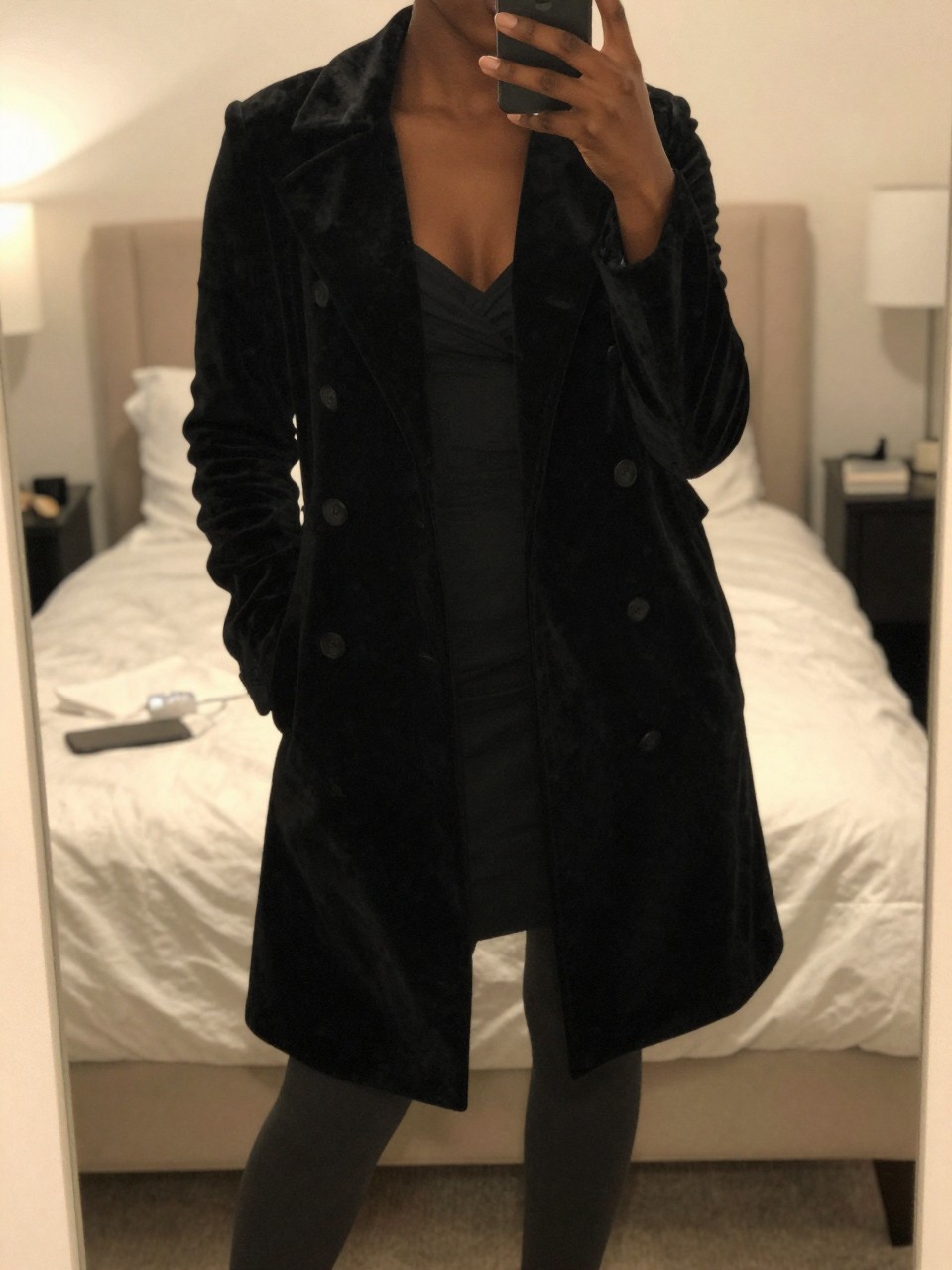 iPhone mirror selfie of a Black woman wearing a black velvet coat over formal evening wear, standing with her phone low and body angled gracefully, cropped at NECK so face doesn't show, in a feminine-themed bedroom, warm moody lighting, iPhone photo quality.
