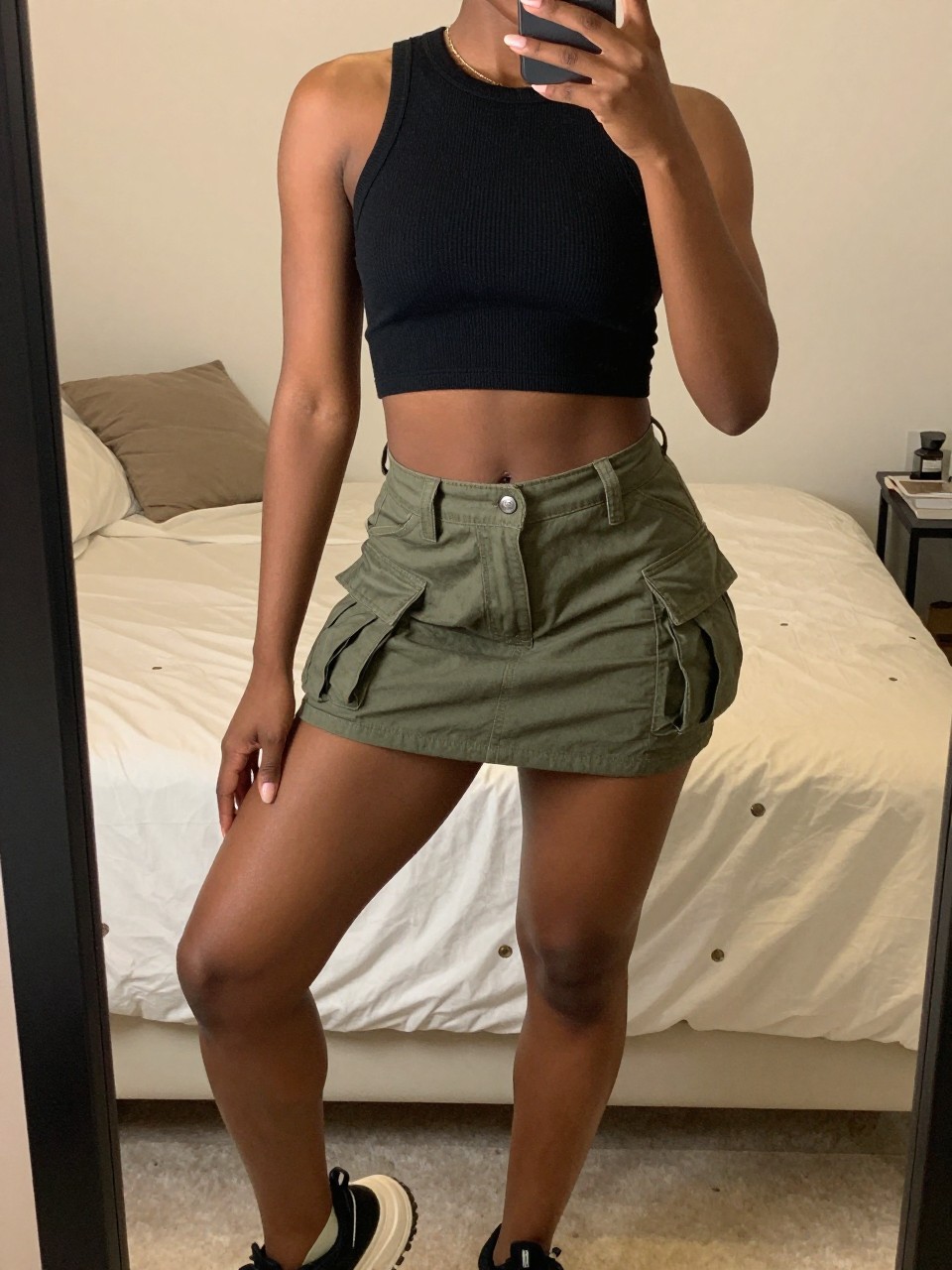 iPhone mirror selfie of a black woman wearing a black ribbed crop top with an olive green cargo mini skirt and chunky platform sneakers, one hand holding her phone low by her thigh, cropped at NECK so face doesn't show, in a feminine-themed bedroom, warm moody lighting, iPhone photo quality.