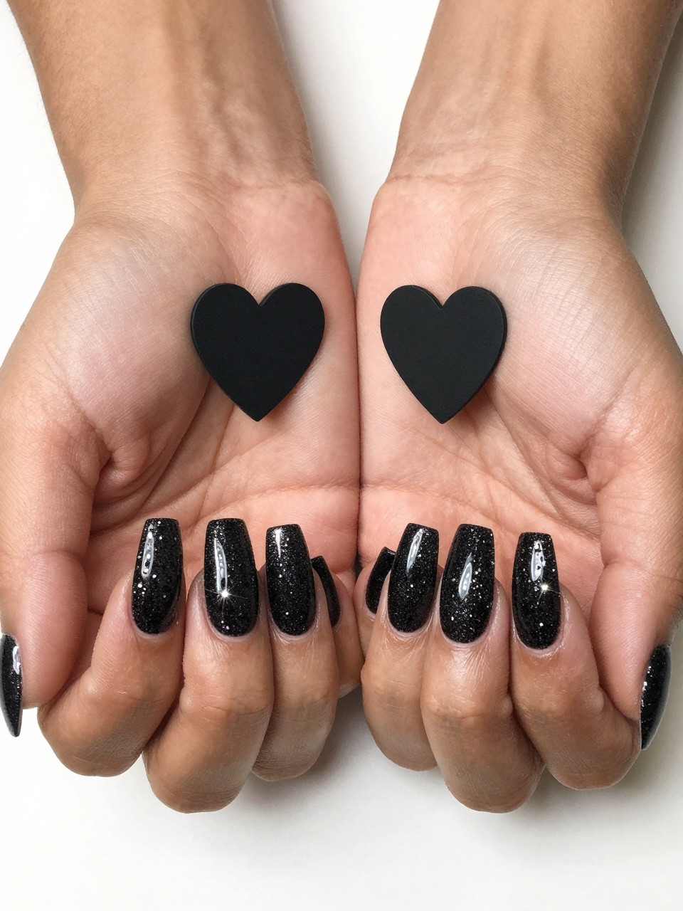 Bird's-eye view of black feminine hands under bright light, showcasing a sparkling black glitter gradient. A solid, flat matte black heart is centered on each nail, absorbing light while the glitter around it explodes with shine.